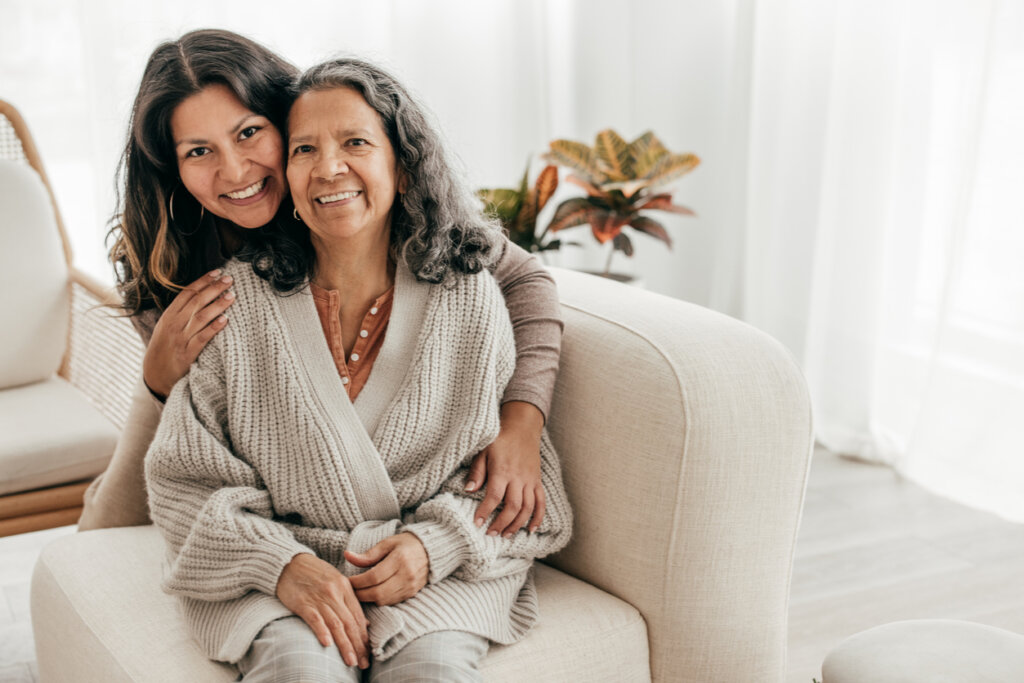 A senior memory care female patient sitting on a couch as her daughter stands behind her with her arms wrapped aorund her shoulders