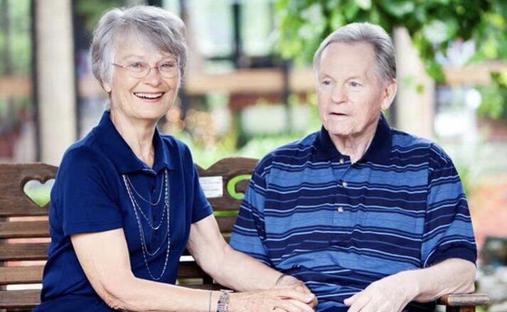 An elderly couple are sitting on a bench outside, smiling and holding hands. Memory care residents enjoy quality time together at Traditions of Grove City.