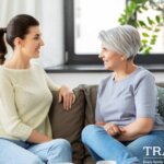 Photo of a woman with dark hair sitting on the couch across from her elderly mother as they have a conversation with the Traditions Management logo in the bottom right corner of the image