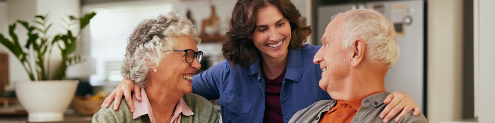 Older couple sitting at a table in a kitchen; a standing woman has her arms draped on the older couple's shoulders. They are all smiling at each other.