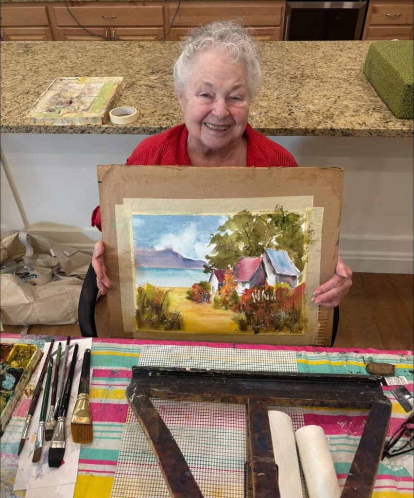 a female Traditions Grove City Senior Living resident smiling while holding a painting in front of a table of arts supplies